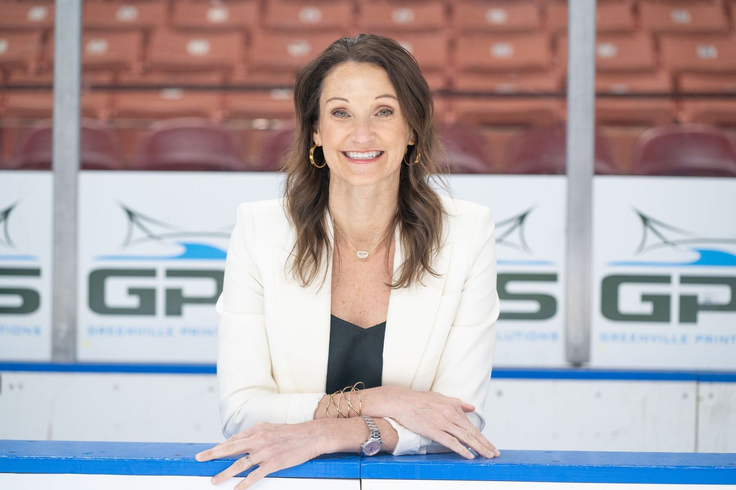 Professional woman in white blazer smiling at desk with sports branding visible in background.
