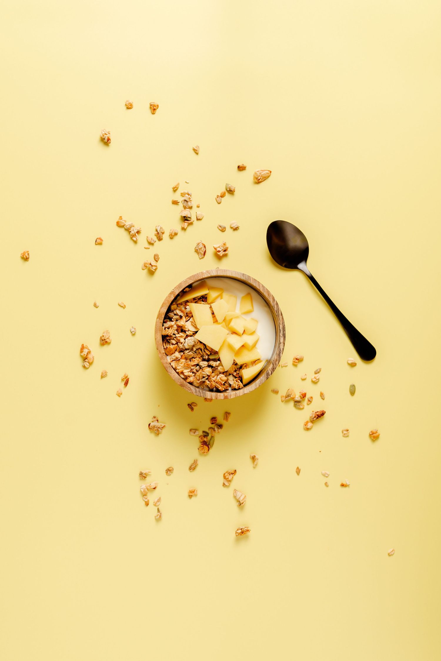 bowl of granola with chopped mango on yellow backdrop with black spoon, healthy eating, lifestyle, vitalising
