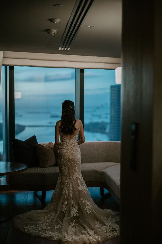 A bride in a lace mermaid gown looks out floor-to-ceiling windows with city views.