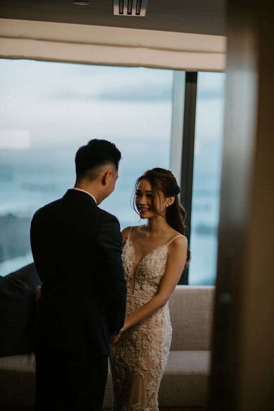 A couple in wedding attire stands together by large windows overlooking a city.
