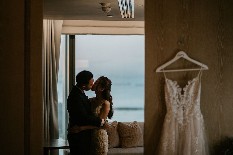 Wedding dress hanging in foreground with couple silhouetted against window in background.
