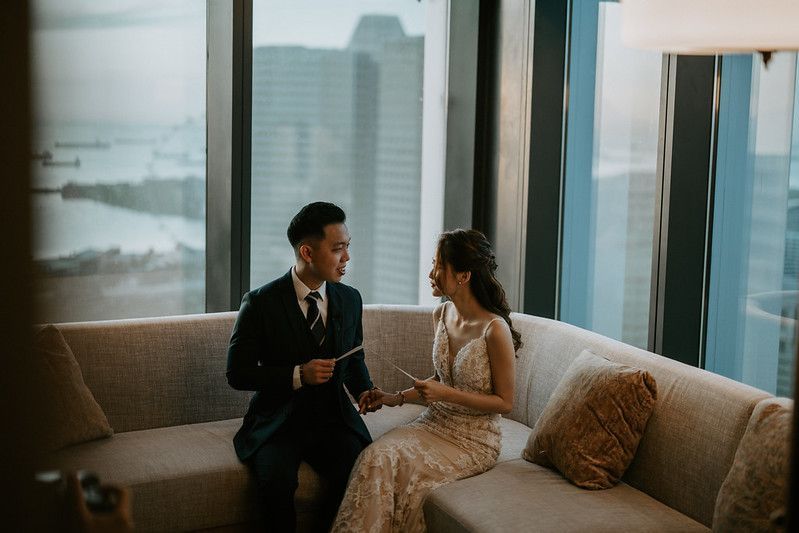 Couple in formal attire sitting close together on grey sofa with cityscape backdrop.