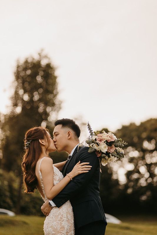 Romantic wedding couple kissing while holding floral bouquet in sunset garden.