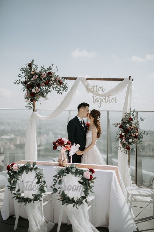Wedding ceremony setup featuring a white fabric arch with pink and red floral arrangements on a city rooftop.