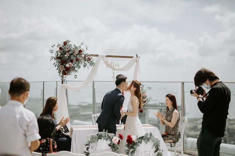Elegant rooftop wedding ceremony with white drapery and red floral accents against cloudy skies.