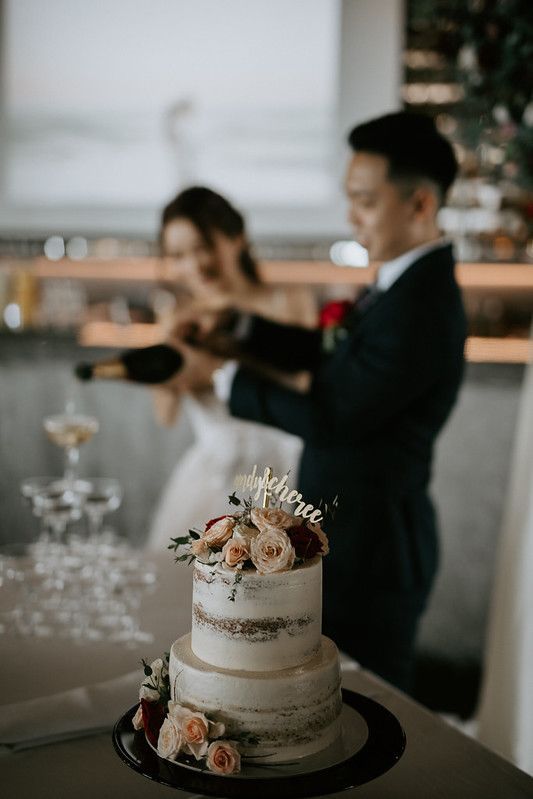 Wedding cake displayed on dark stand with champagne tower setup in background.