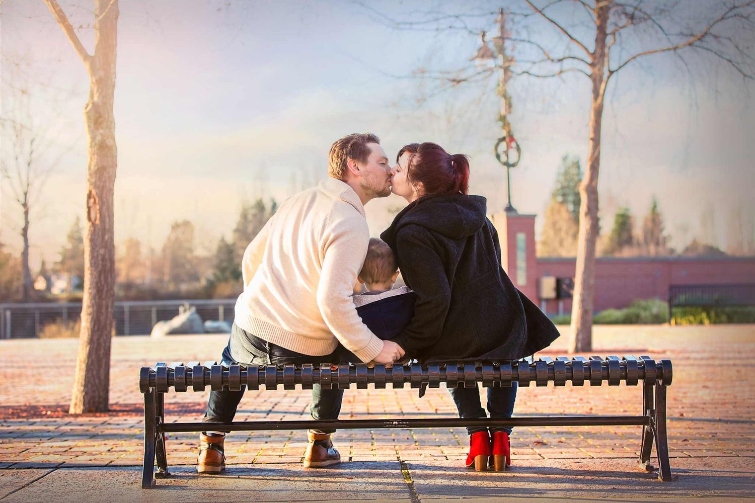 Family of three sitting facing away on a bench together. Parents are kissing over the toddlers head at sunset.