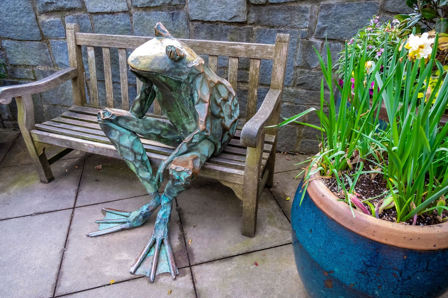 Bronze frog sculpture sitting on wooden garden bench next to potted plants.
