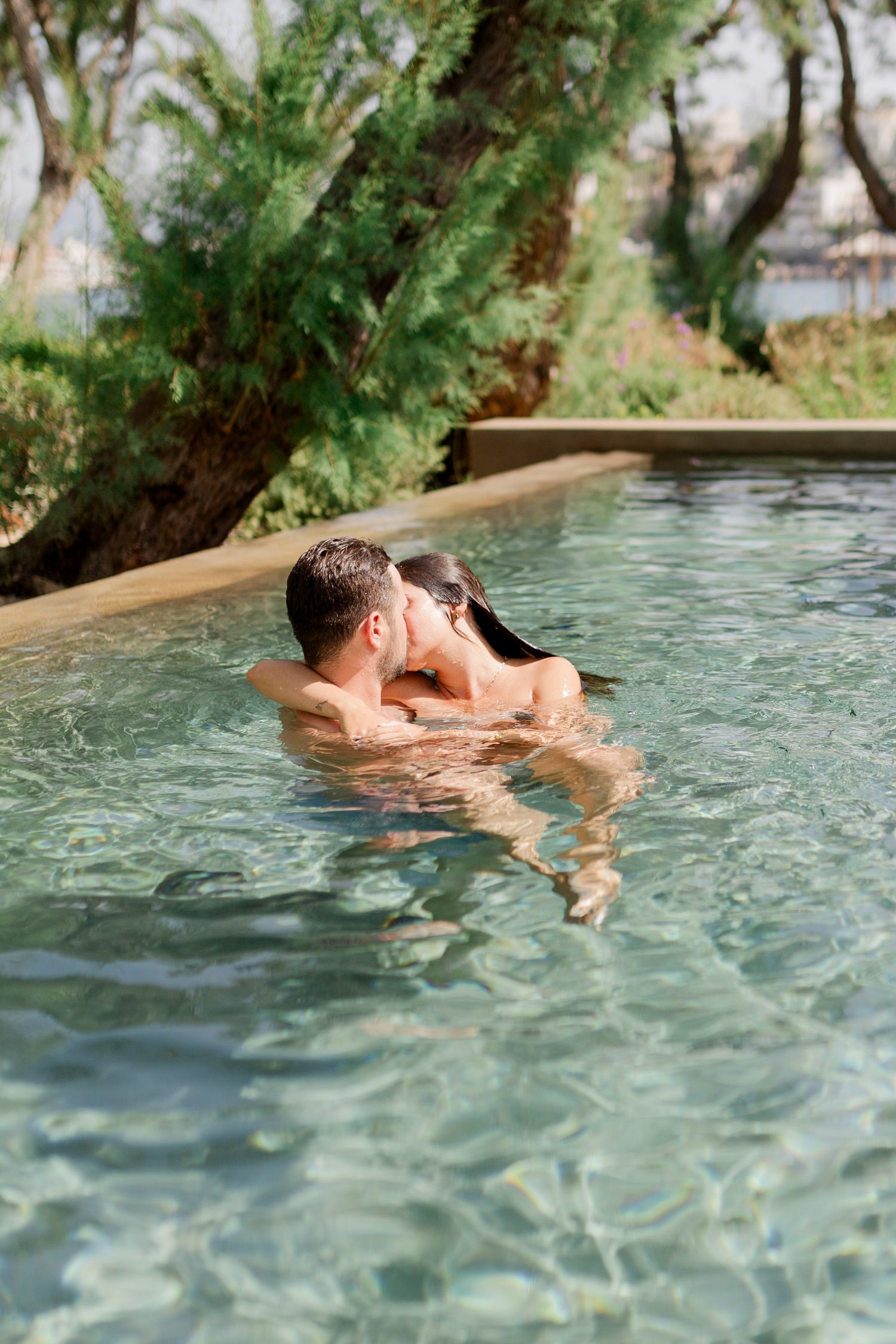 Two people embracing in a swimming pool surrounded by lush greenery.