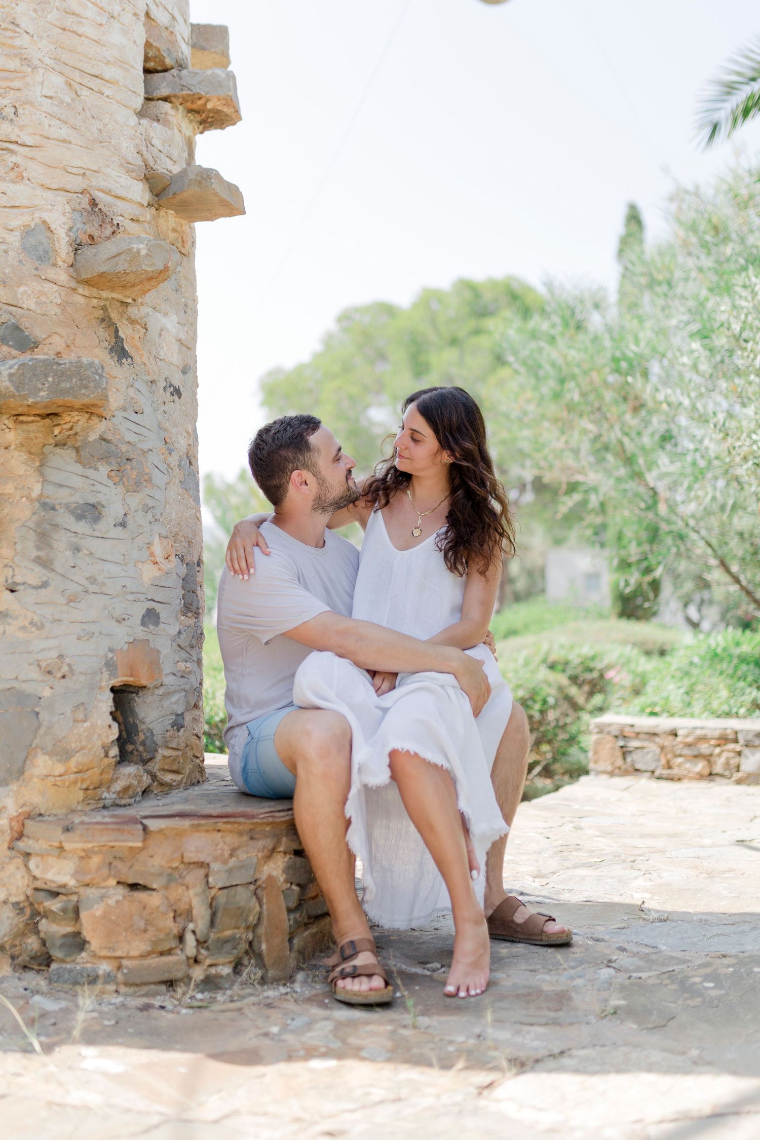A romantic couple sitting on stone steps by an ancient windmill wall in summer attire.