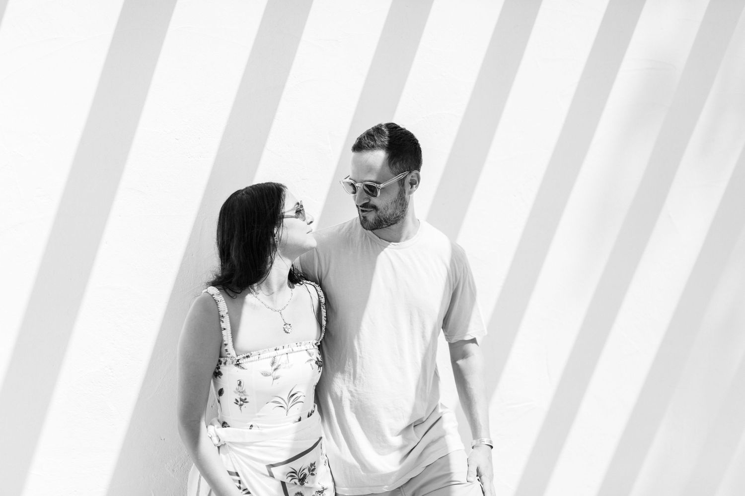 Couple standing close together against white wall with striped shadows.