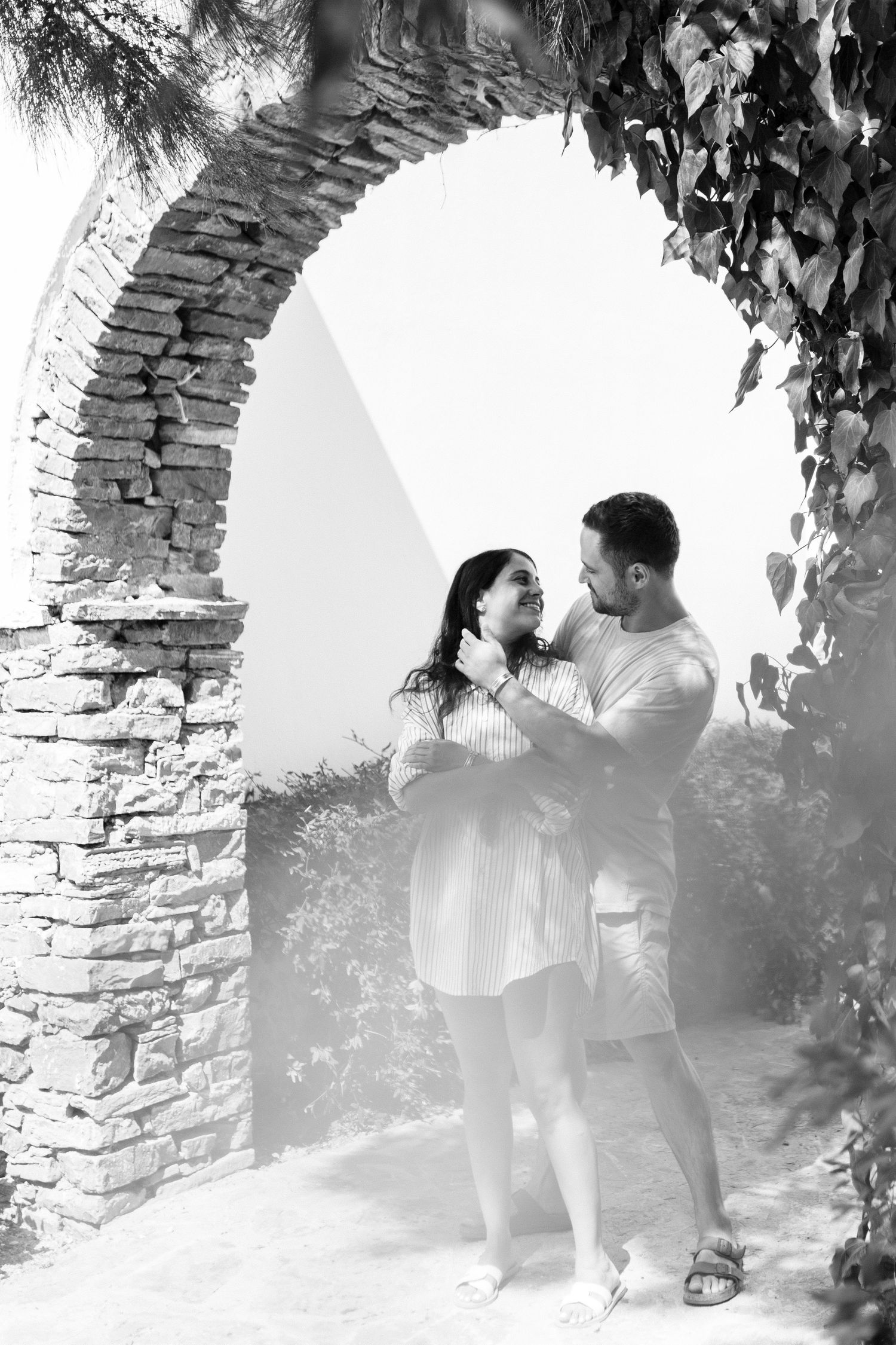 A black and white photo of a couple embracing under a rustic stone archway.