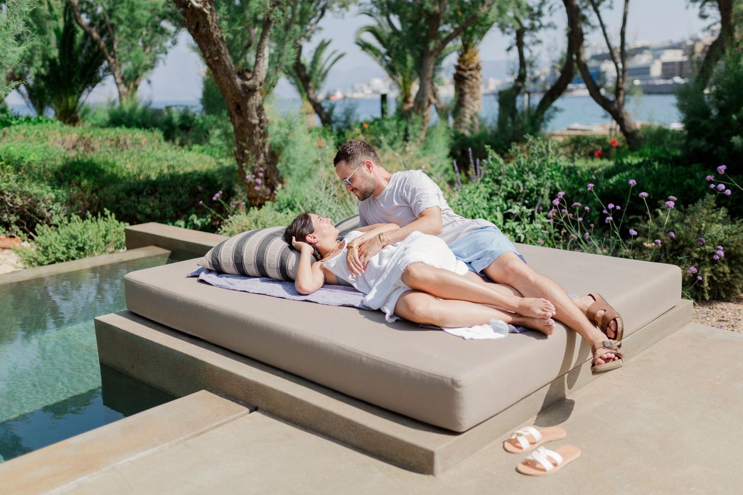 A couple relaxes together on a cushioned lounge chair next to a pool surrounded by lush greenery.