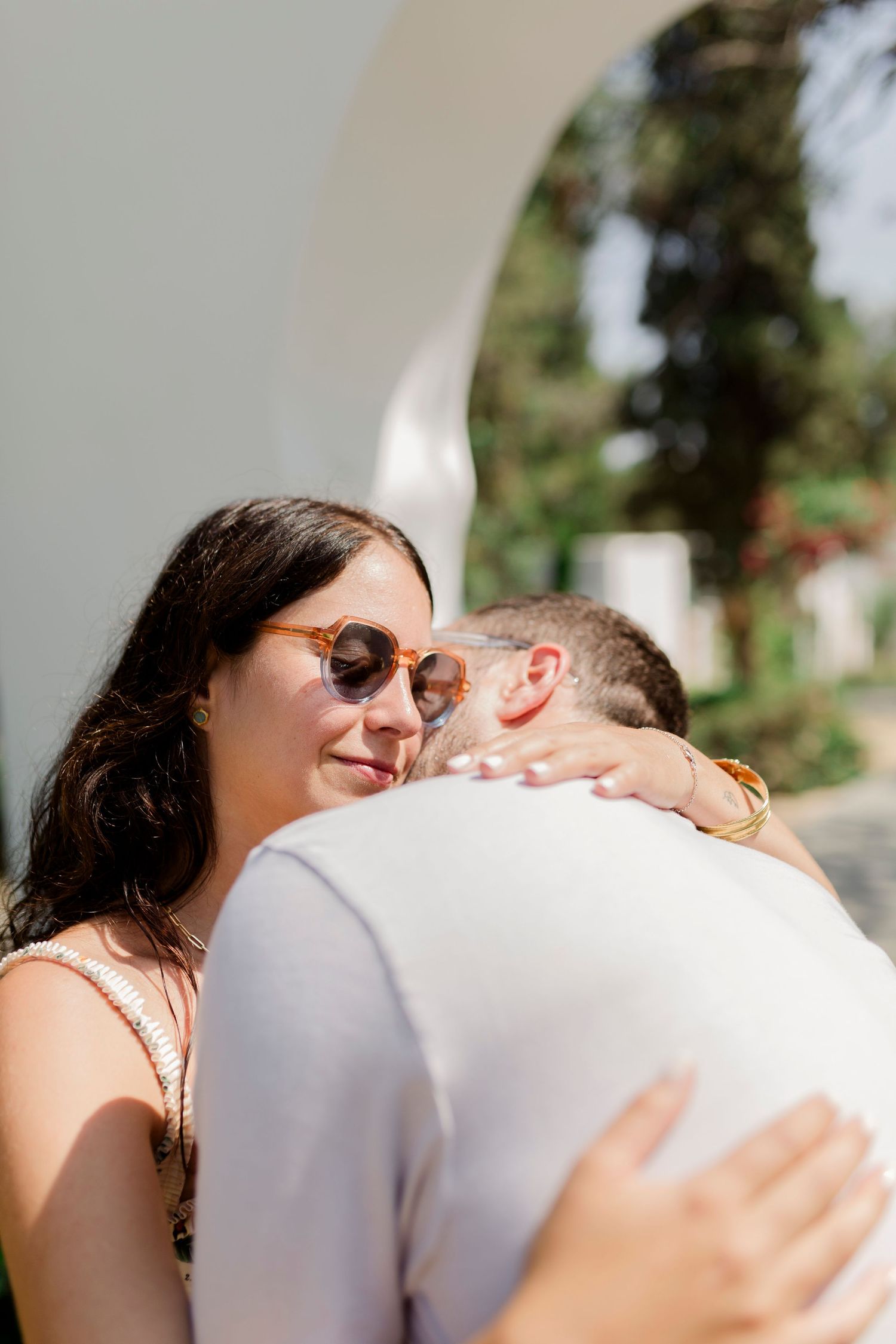 A couple embraces under a white archway, surrounded by lush greenery and trees.