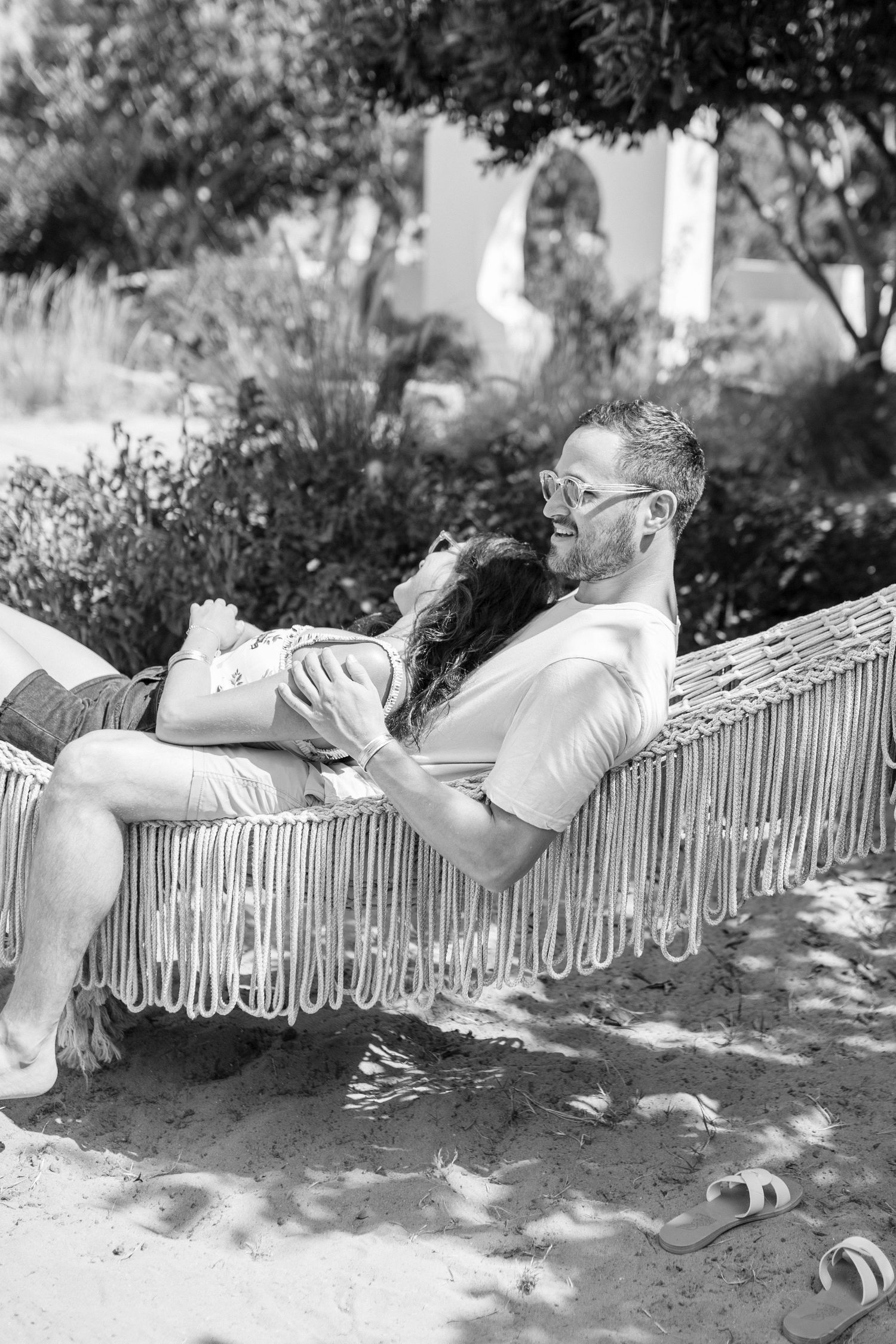 A couple reclines in a macrame hammock in a garden, with the man cradling his partner.