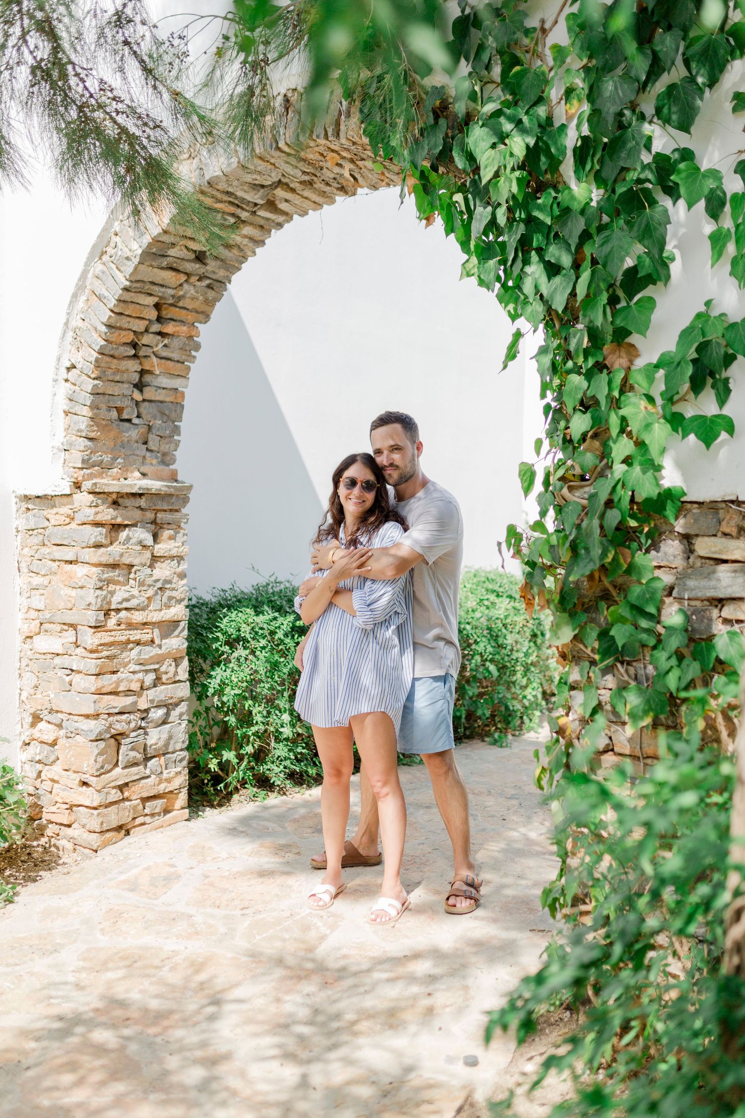 A couple sharing a tender moment under a vine-covered stone arch in summer clothing.