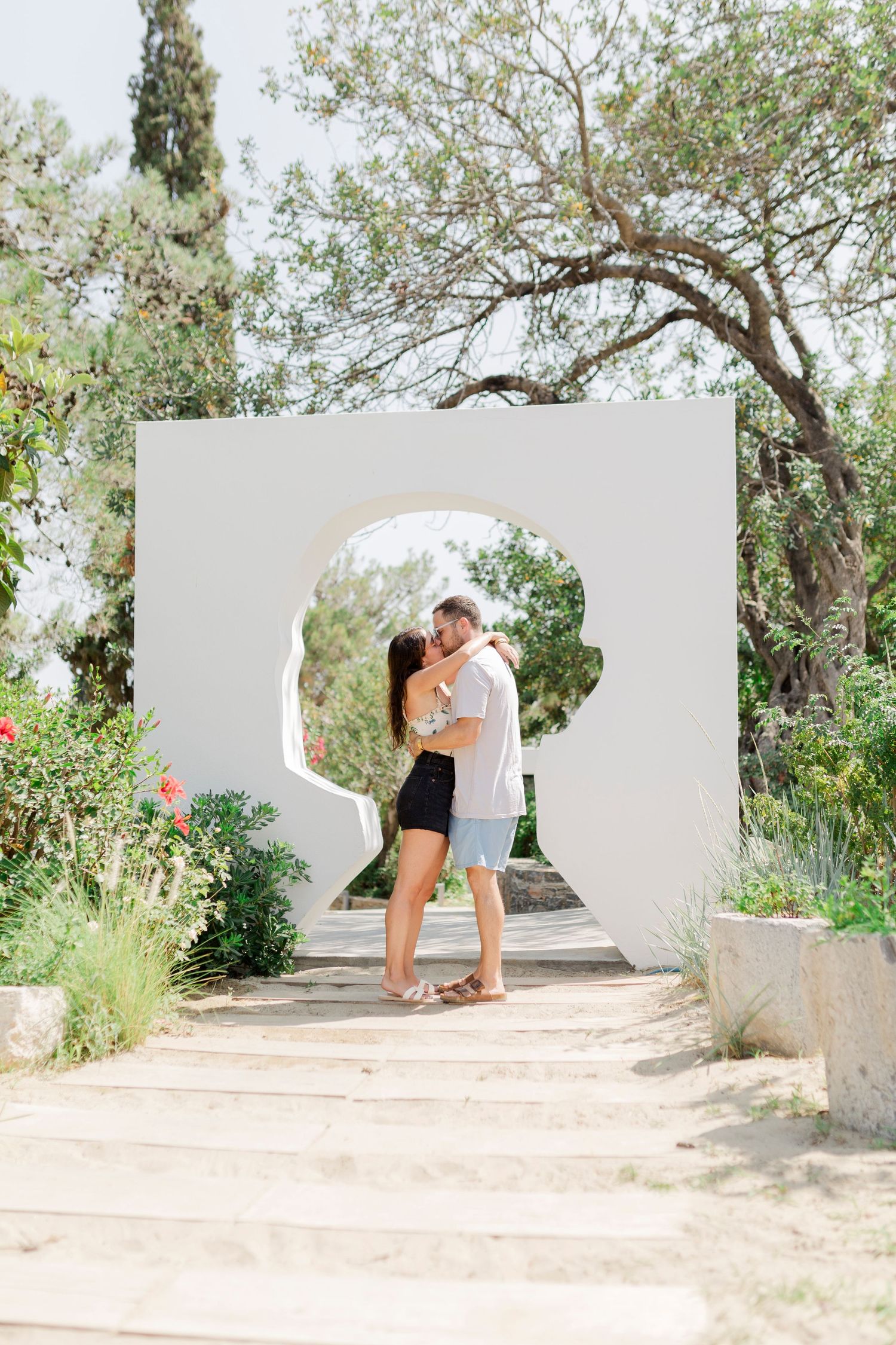A couple stands in an arched white doorway, surrounded by greenery and trees.