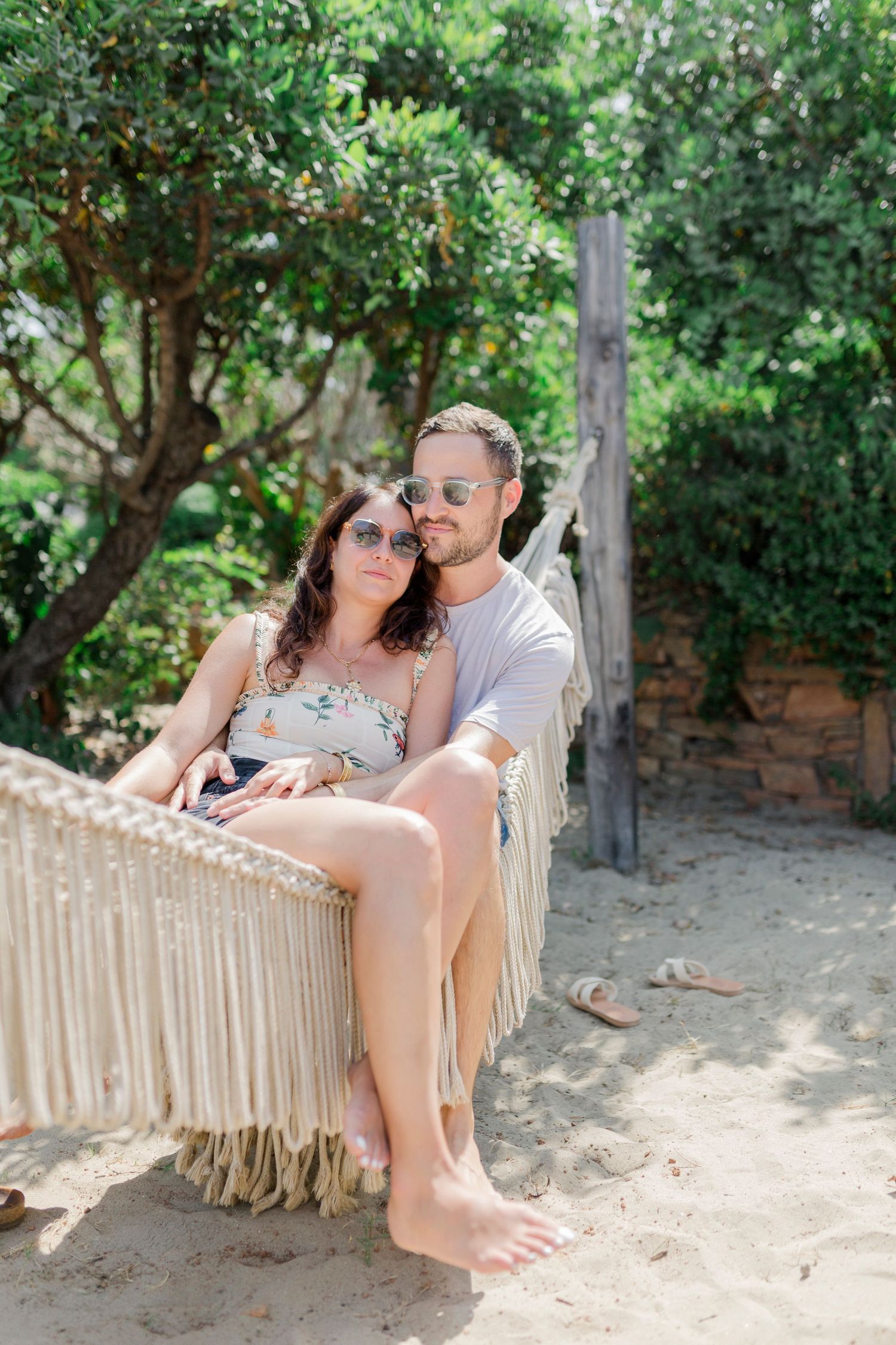 A couple lounges in a fringed hammock in a sunny garden setting with trees and foliage.