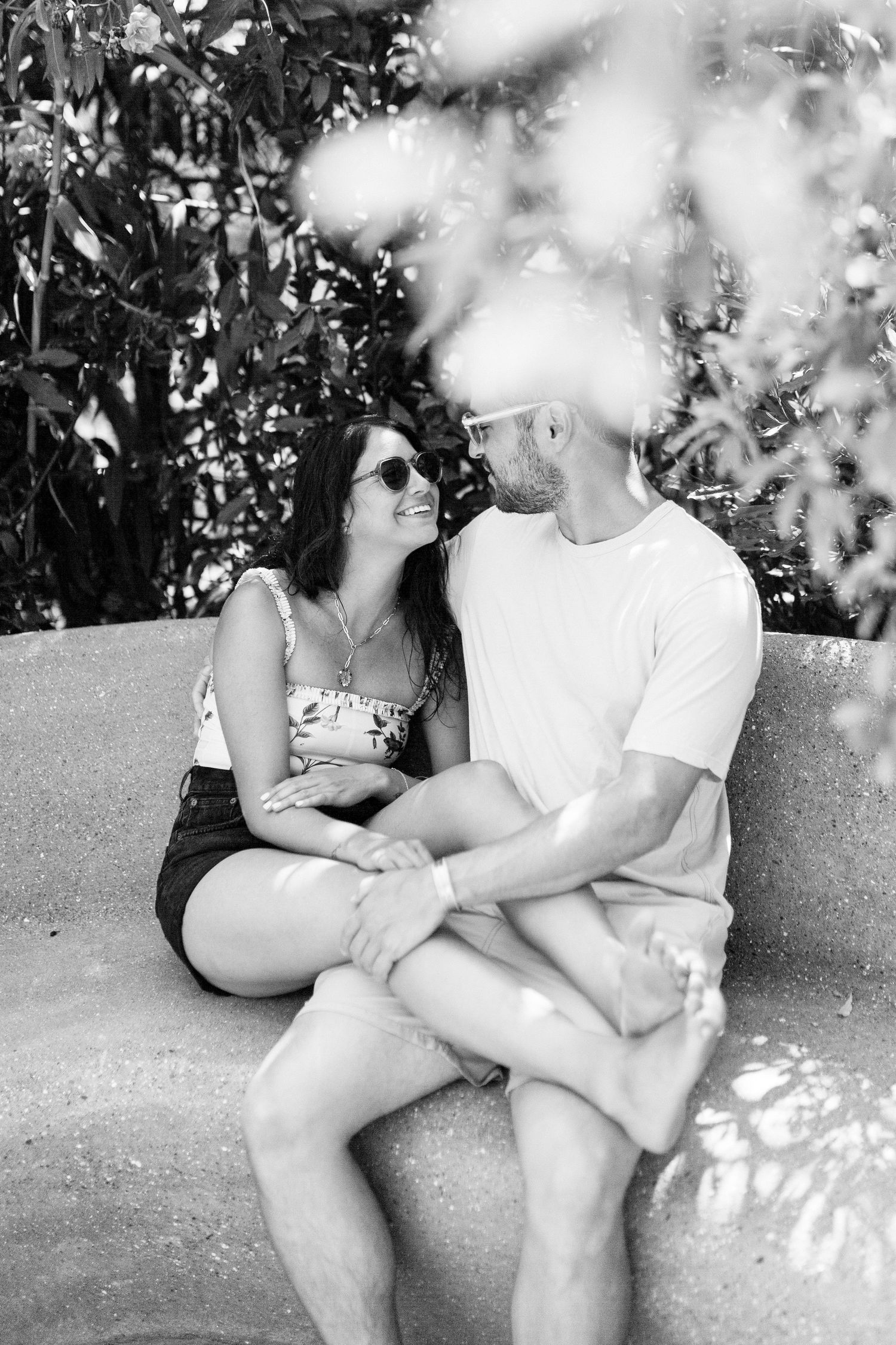 Couple sitting close together on concrete surface surrounded by foliage.