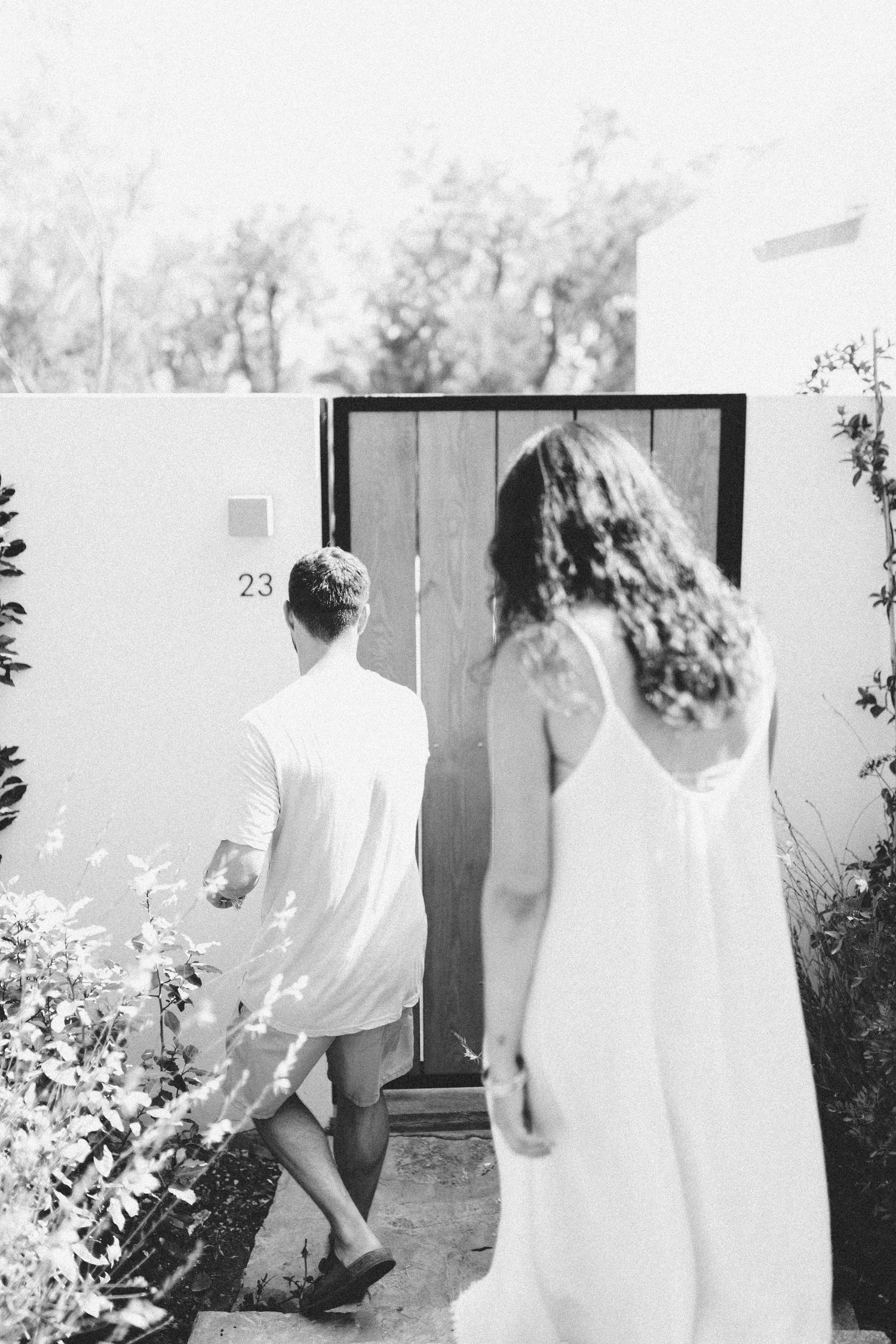 A couple in white clothing walking towards a wooden door in a Mediterranean garden setting.