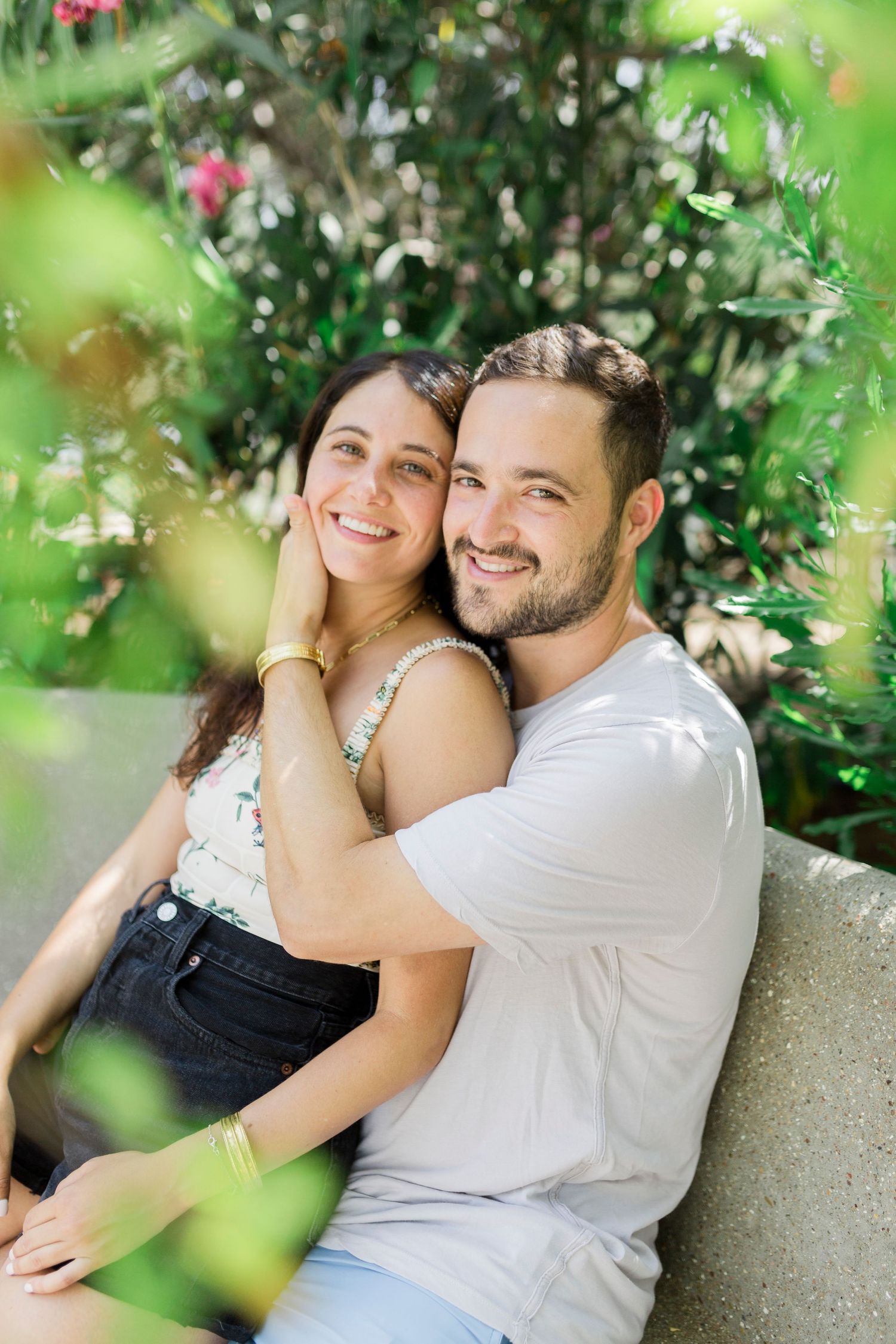 A smiling couple embraces in front of greenery and flowers in a garden setting.
