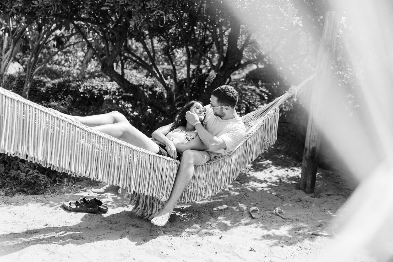A couple embraces while resting in a fringed hammock in a garden, captured in black and white.