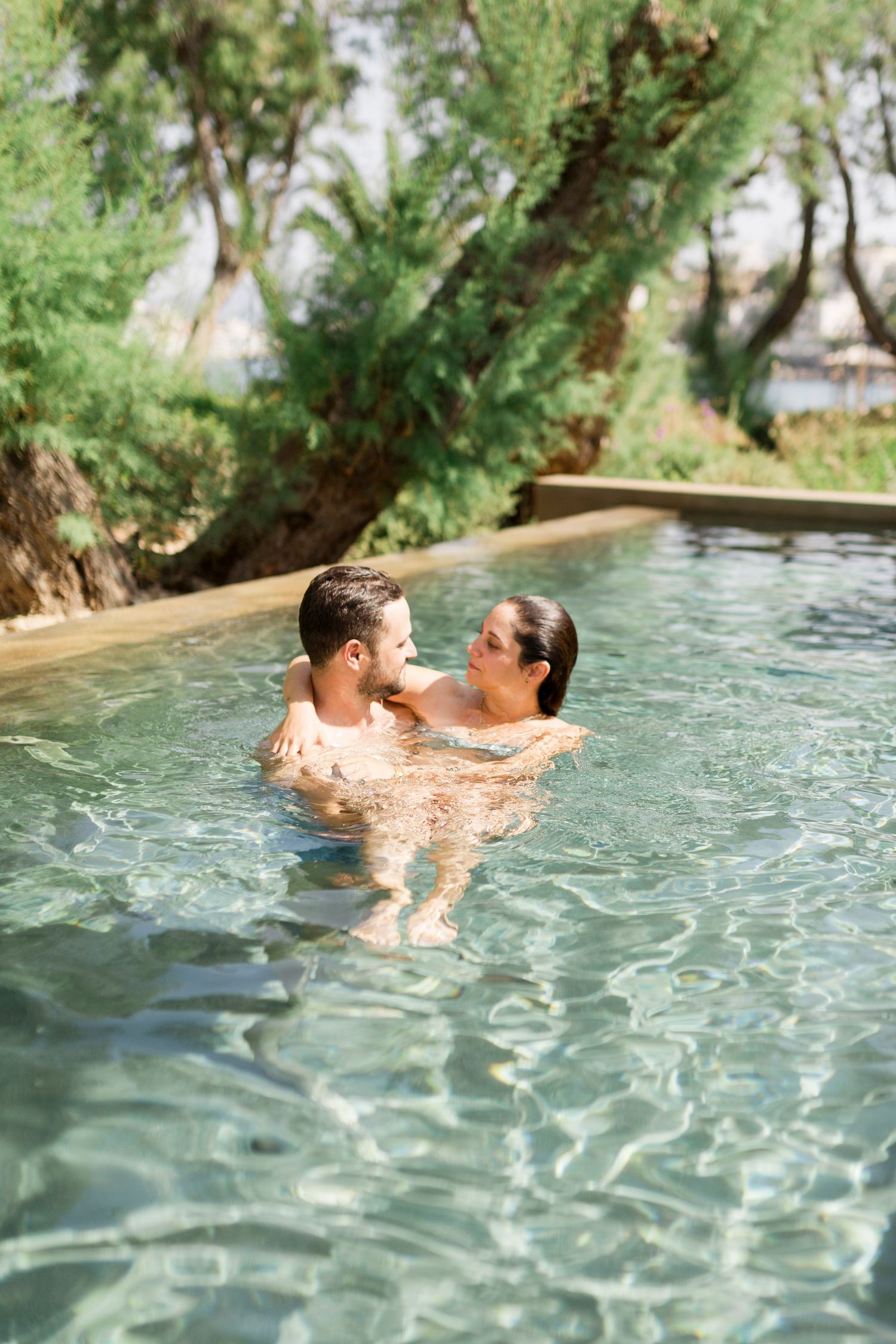 Couple embracing in a swimming pool surrounded by trees and plants.