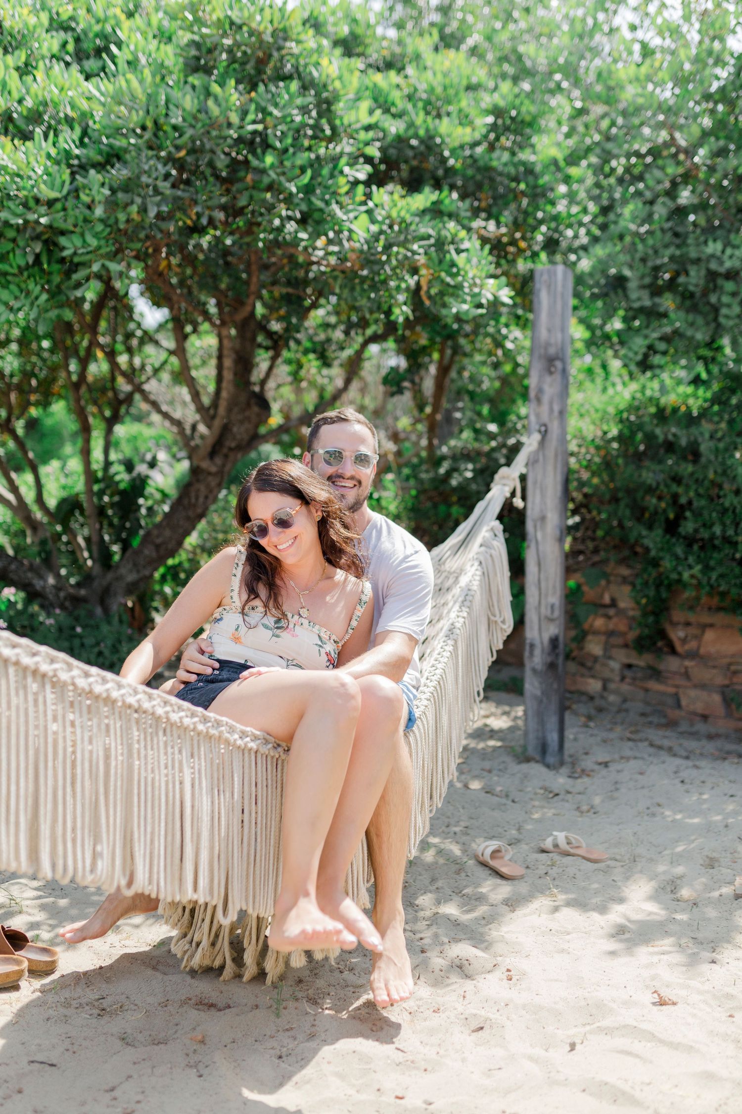 A couple sits together in a fringed hammock surrounded by greenery and sandy ground.
