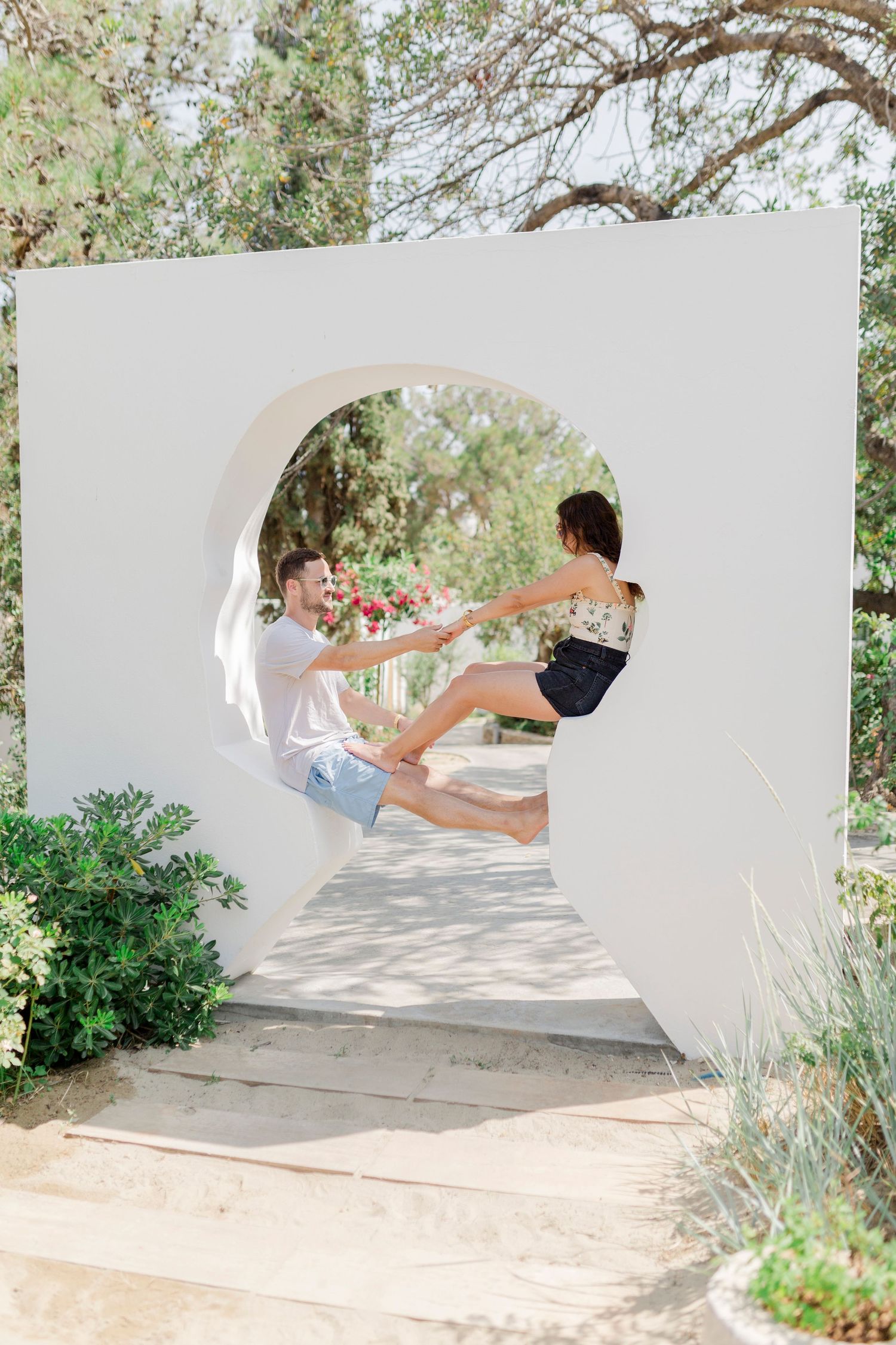 A couple sits playfully in a circular white archway surrounded by lush greenery and trees.
