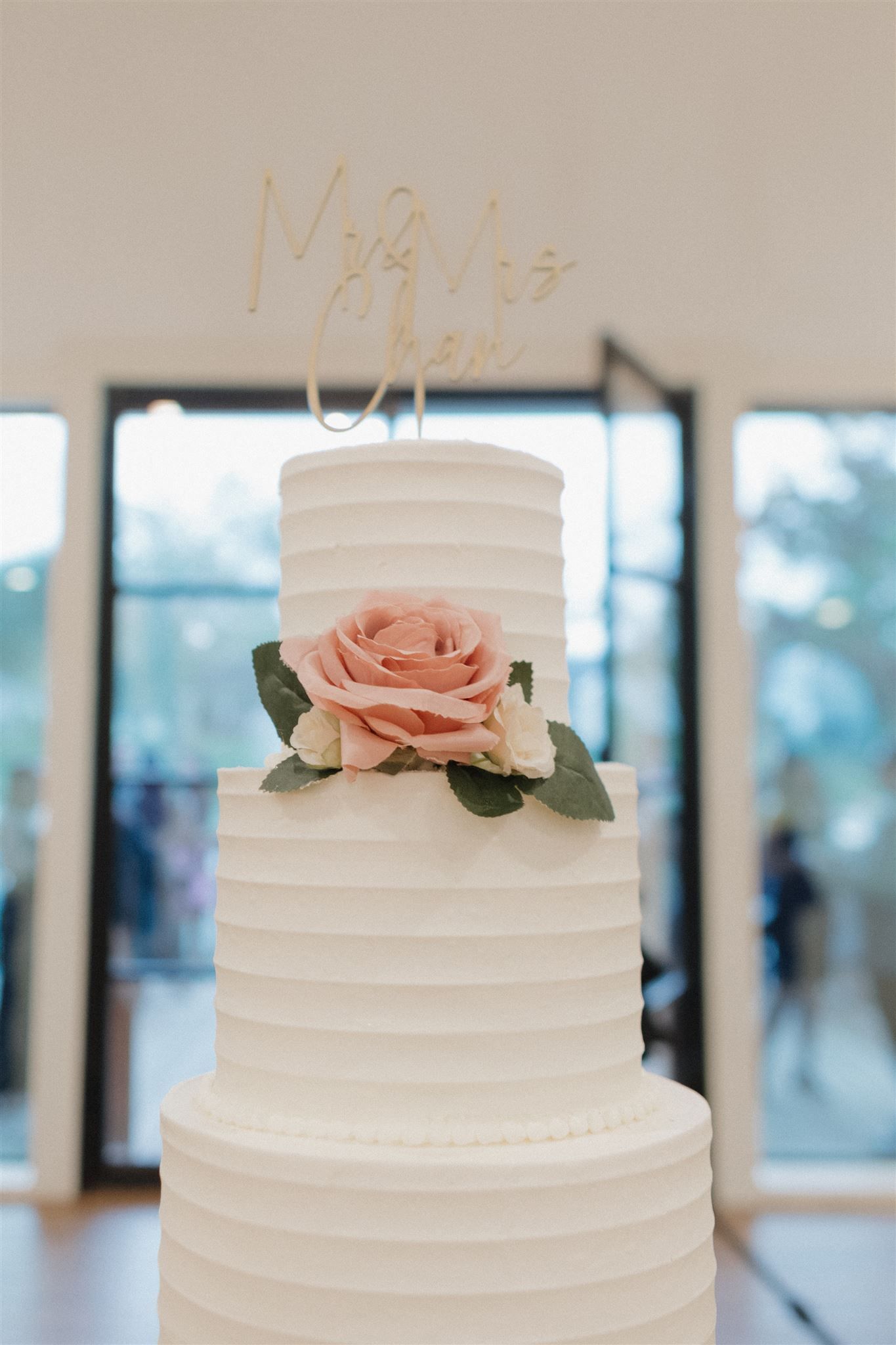Three-tiered white wedding cake with blush pink rose and gold 'We Do' topper.
