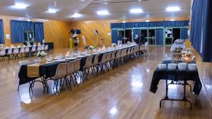 Long banquet tables arranged in rows with white chairs in a well-lit gymnasium.