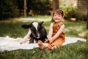 Child in brown overalls sitting happily next to black and white goat on blanket.