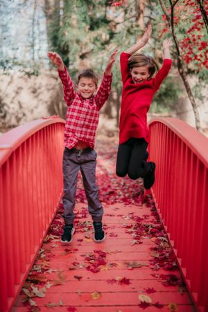 Two children jumping with joy on red bridge covered in fallen autumn leaves.