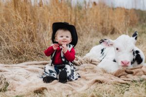 A small child in western wear sits on blanket with spotted calf among tall grasses.