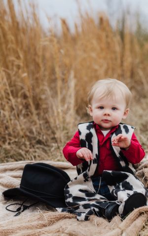 A toddler dressed in cow print vest and red shirt sits with a black cowboy hat on a blanket.