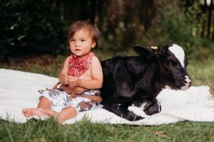 Child in red bandana sits next to resting black and white baby goat on white blanket.
