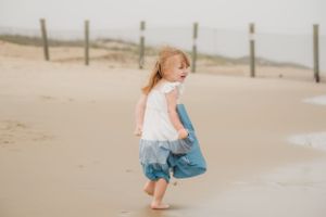 A child wearing a white and blue dress dances barefoot along the sandy shoreline.