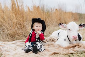 A baby in black cowboy hat sits beside a curious white calf on outdoor blanket.