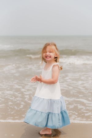 A happy child plays at the beach wearing a white and blue tiered dress with waves in the background.