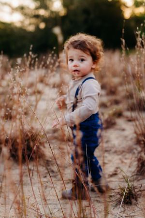Small child in suspenders standing among tall dried grass during sunset.