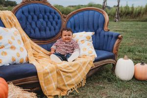 Child sits on blue vintage sofa decorated with yellow throw and pumpkin pillows next to autumn gourds.