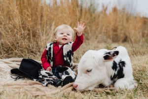 A small child in western wear waves while sitting with a spotted goat on a blanket.