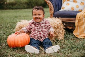 Smiling child in plaid shirt sits by hay bale with large orange pumpkin on grassy field.