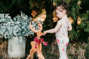 Child in floral outfit plays with a toy reindeer decoration near potted plants.