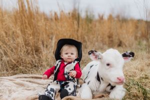 A baby in western attire sits next to a white spotted calf on a blanket outdoors.