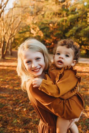 Two children in matching brown outfits share a playful moment among autumn trees and fallen leaves.