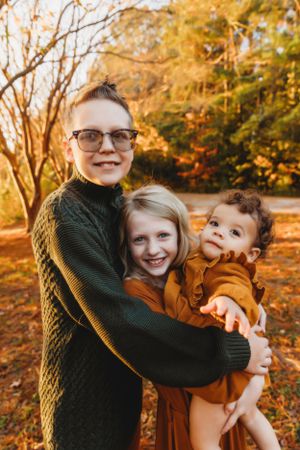 Three siblings embrace in a warm hug while standing in a sunlit autumn forest setting.