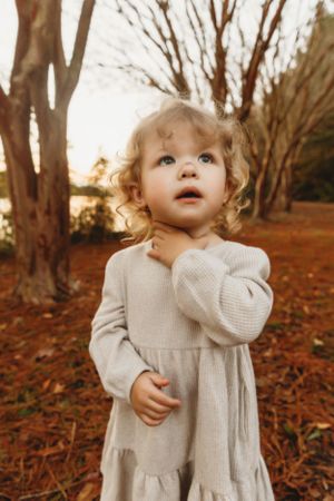 A small child in a cream-colored dress looks up with wonder among bare autumn trees.