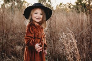 A small child wearing a black hat and rust-colored dress stands joyfully among tall autumn grass.