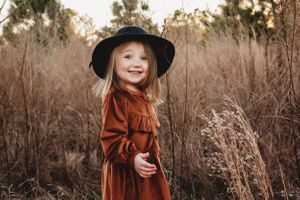 A young child in a brown dress and black hat poses in tall autumn grass at sunset.
